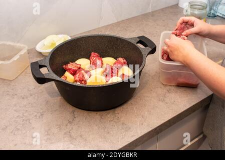 Frau Hände Bereiten Koch Rindfleisch Auf Küche. Nahaufnahme des Kochens Traditionelles jüdisches Cholent Hamin Hauptgericht für den Shabbat.Rohes Rindfleisch wird einem Topf mit Kartoffeln, Zwiebeln, Bohnen und Weizenkörnern hinzugefügt Stockfoto