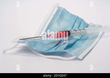 Eine Spritze, gefüllt mit roter Flüssigkeit, wie Blut, auf einer chirurgischen Maske, symbolisch für Impfung und medizinische Behandlung Stockfoto