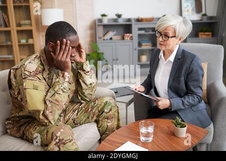 Deprimierter Militärmann sitzt auf dem Sofa und hält seinen Kopf mit Hände und Gespräch mit Psychologen während des Treffens Stockfoto