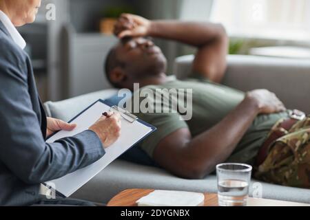 Nahaufnahme einer Frau, die ein Rezept in ihrem Dokument mit schreibt Ihre Patientin liegt auf dem Sofa im Hintergrund Stockfoto
