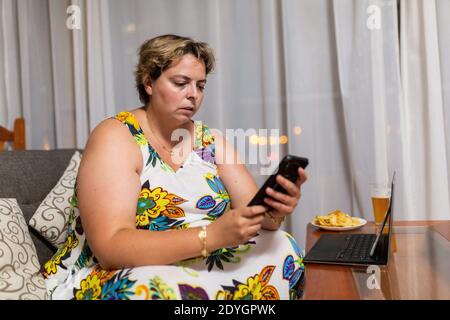 Übergewichtige Frau in einem Blumenkleid auf dem Sofa sitzend mit einem Handy in den Händen, Laptop auf dem Tisch mit einem Teller Chips und ein Glas Bier Stockfoto