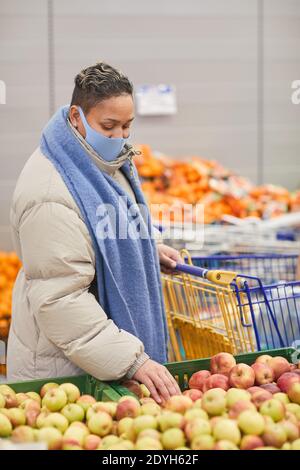 Junge Frau in warmer Kleidung und in Maske Auswahl frisch Obst im Supermarkt Stockfoto