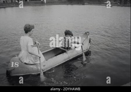 1950s, historisch, eine junge Mutter in einem ärmellosen Kleid sitzt auf der Rückseite eines Kanus, mit ihrem jungen Sohn im Voraus, zusammen paddeln auf einem großen See Boot, England, Großbritannien. Und in dieser sorglosen Ära, in der Gesundheit und Sicherheit nicht die Branche war, die es wurde, keine Rettungsweste oder Hi-vis-Top vor Ort. Stockfoto