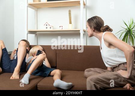 Mama schimpft die Söhne, die auf der Couch liegen und herumliegen. Erziehung von Söhnen im Teenageralter. Mama schimpft Kinder. Stockfoto