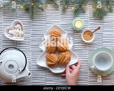 Kreative Xmas Tischaufstellung für Tee. Draufsicht mit Eclare, Kuchen backen. Hand hält Teller mit Kuchen in Form von Weihnachtsbaum. Tannenzweige mit Licht Stockfoto