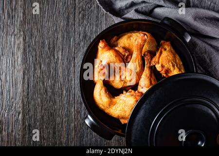 Knusprig geröstete Hähnchenbeine in schwarzer Keramikpfanne auf einem dunklen Holztisch, horizontale Ansicht von oben, flach liegend, freier Platz Stockfoto