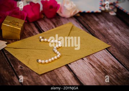 Geschlossener Umschlag mit einer Perlenkette auf der Oberseite und einem Geschenkbox im Hintergrund mit Rosen und alten Buchstaben Stockfoto