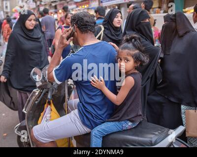 Umgeben von muslimischen Frauen in Burqas hält ein Vater sein Motorrad an, um einen Anruf zu tätigen, seine Tochter klammert sich auf dem Rücksitz an ihn, Mumbai, Indien Stockfoto