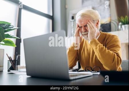 Älterer müder grauhaariger Mann, der seinen Kopf hält und seine Augen vor Kopfschmerzen schließt, ist er müde von langer Arbeit in einem Laptop Stockfoto