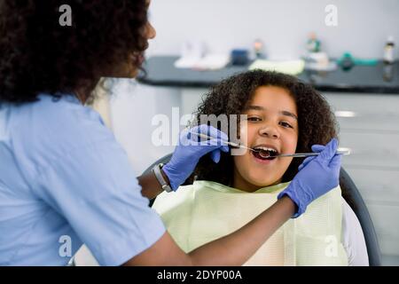 Close up Draufsicht auf hübsches Kind, afroamerikanische lockige Mädchen in Zahnarztstuhl, mit offenem Mund, mit Zahnuntersuchung. Rückansicht der weiblichen Afrikanerin Stockfoto