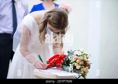 Eine Braut, die eine medizinische Maske mit einem Blumenstrauß trägt, unterschreibt während der Coronavirus-Panademie eine Heiratsurkunde. Stockfoto