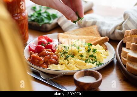 Servieren Sie Rühreier mit Brot Toast und gebackenen Bohnen. Frau garniert Frühstück Eier Omelette mit gehackter Petersilie Stockfoto