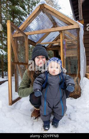 Mama und Kleinkind posieren für die Kamera kniet im Schnee während der Erkundung eines Hühnerstall im Schnee. Die ersten Entdeckungen des Babys. Stockfoto