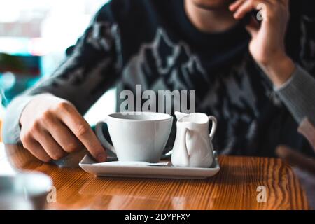 Mann, der in einem Café telefoniert, mit einer Kaffeetasse und einer Milchkaffee auf dem Tisch. Hand- und Winterpullover für Erwachsene Stockfoto
