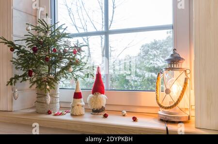 Weihnachtslaterne, Weihnachtsknome, Weihnachtsbaum und rotes Dekor auf dem Fenster eines Holzhauses mit Blick auf den Wintergarten. Stockfoto