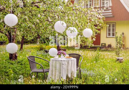 Terrassenmöbel im Apfelgarten auf wildem, langen Rasen, Tisch mit weißer Tischdecke und weißen Papierlaternen, die vom Apfelbaum hängen. Niedliches Holz. Stockfoto