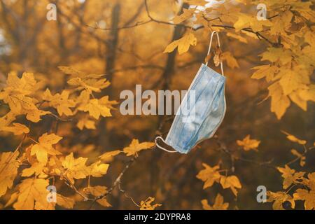 Eine gebrauchte Gesichtsmaske, die in herbstfarbenen Baumzweigen in einem Stadtpark hängt. Öffentliche Räume werden mit ausrangierten Schutzausrüstungen überflutet. Stockfoto