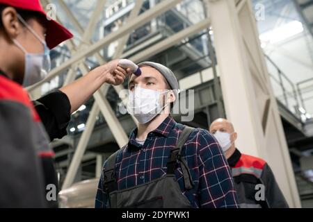 Junge Fabrikarbeiterin in Schutzmaske zur Messung der Körpertemperatur Von einem der Ingenieure gegen riesige Konstruktion Stockfoto