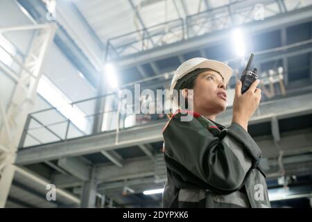 Junge Ingenieurin in Schutzhelm und Arbeitskleidung mit Walkie Talkie, während in der großen Werkstatt der zeitgenössischen Fabrik stehen Stockfoto