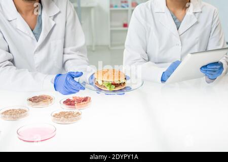 Handschuhen des Arbeiters der Lebensmittelqualitätskontrolle, die Platte hält Mit Hamburger, der Gemüsefleisch enthält, während sein Kollege Daten eingibt Stockfoto