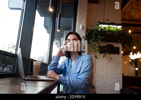 Seriöse Geschäftsfrau, die am Telefon spricht und auf die Kamera schaut. Seitenansicht. Gemütliche Kaffeehaus-Atmosphäre. Freiberufler mit modernem Laptop für Stockfoto