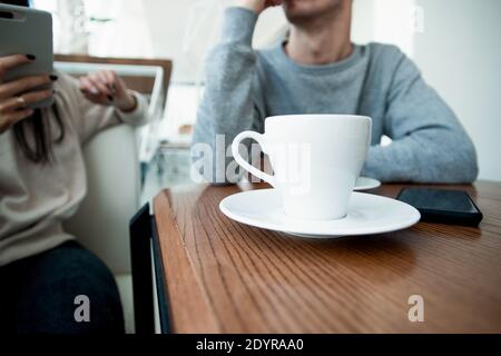 Weiße Tasse Kaffee auf der Vorderseite. Verschwommenes Paar auf dem Hintergrund. Mann und Frau auf romantische Datum im Café. Kaffeehaus. Frau hält ihr Smartphone und Stockfoto