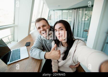 Fröhliches Paar, das Selfie nimmt. Mann umarmt seine Freundin und humbing up. Frau holdig Smartphone und Blick auf die Kamera. Heller Raum im Hintergrund Stockfoto