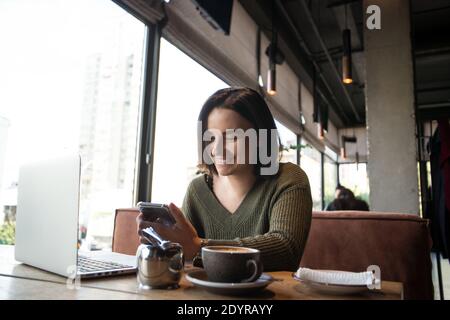 Glückliche junge Frau im Café lächelt Blick in Smartphone. Konzept der Online-Kommunikation. Hübsche junge Frau sitzt in einem Café mit Laptop geöffnet Stockfoto