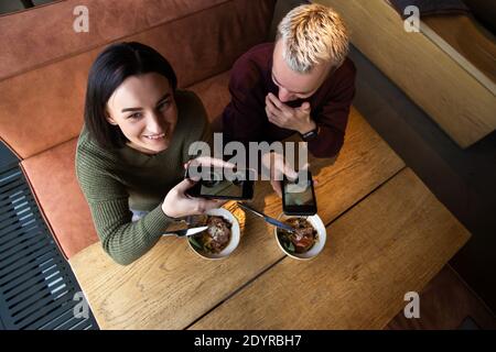 Paar fotografieren von Essen. Brunette Frau hält Smartphone, wegschauen und lächeln. Blonde Mann fotografieren und lachen. Gute Laune Konzept Stockfoto