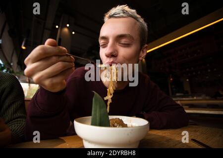 Zufriedener Mann genießt das Essen im Restaurant. Blong attraktiver junger Mann mit durchbohrter Nase setzt Pasta in den Mund und schließt die Augen mit Vergnügen Stockfoto