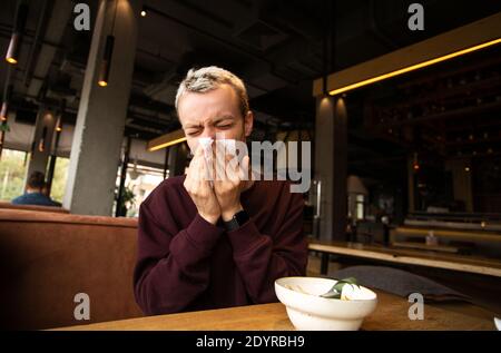 Kranker Mann mit laufender Nase kam in ein Café. Bleib zu Hause Konzept. Blonde junge Mann bläst seine Nase in öffentlichen Ort. Isolieren Sie sich selbst, wenn Sie krank werden Stockfoto