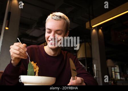 Gutes Kundenservicekonzept. Glücklicher junger Mann lächelt und zeigt seine Zähne und schaut auf leckeres Essen auf seiner Gabel. Gut aussehende blonde Mann in legerer Kleidung Stockfoto