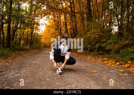 Junge Frau Trailrunner binden Schnürsenkel im Wald. Gemütliche Herbstsaison im Freien. Gesundes Lifestyle-Konzept. Herbstlaub auf der Straße. Nette Frau mit Stockfoto