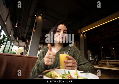 Leckeres Essen im Restaurant. Fröhliche Brünette Frau zeigt Daumen nach oben Geste mit einer Hand und hält ein Glas Orangensaft in einer anderen. Schüssel mit Stockfoto