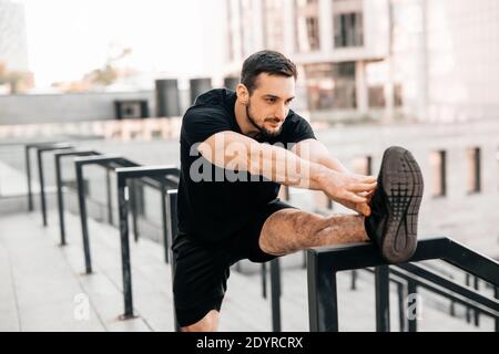 Mann, der sich vor der morgendlichen Aktivität zurückstreckt. Sport im Großstadtkonzept. Athletischer Mann, der Hände bis zum Bein streckt. Konzentrierter Läufer, der sich vorher aufwärmt Stockfoto