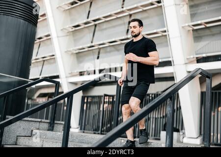 Athletischer junger Mann, der von der Treppe herunterläuft. Urbanes Sportkonzept. Apartmentblock im Hintergrund. Aktives Leben. Fitness im Freien. Mann in Schwarz Stockfoto