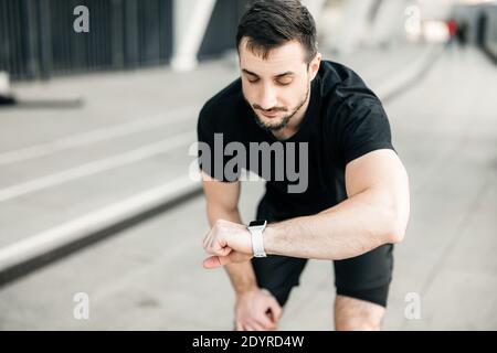 Der Läufer ruht sich aus und schaut auf seine Uhr. Cardio Load Control Konzept. Attraktive Brünette Mann Joggen am Morgen in schwarzen Sportkleidung. Grau Stockfoto