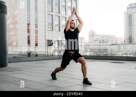 Der sportliche Mann übt die Low-Ausfallschritt-Pose mit erhobenen Armen. Junger Mann in schwarzem Sport-T-Shirt und Leggins Stretching in Yoga-Pose. Urbanes Sportkonzept Stockfoto