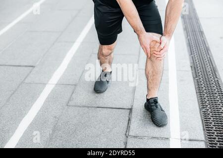 Männliche Hände halten schmerzende Knie. Starker Mann, der beim Joggen im Freien Schmerzen im Fuß spürt. Nahaufnahme. Männlicher Läufer in schwarzen Sneakers stehend und Stockfoto