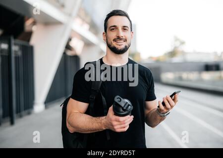 Fröhlicher Mann, der die Kamera anschaut und lächelt. Junger männlicher Athlet in schwarzer Kleidung mit Sportflasche und Smartphone in den Händen. Attraktiver Mann ist Stockfoto
