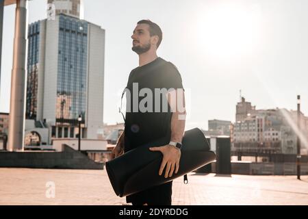 Selbstbewusster junger Mann hält gerollte Yoga-Matte und Blick auf schöne Morgen Blick auf die große Stadt. Konzept der morgendlichen Aktivitäten. Strahlender Sonnenschein Stockfoto