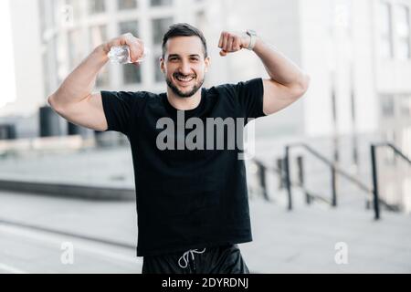 Fröhlicher Mann, der seine Muskeln zeigt. Hartes Trainingskonzept. Fit Mann zeigt Bizeps und lächelt. Graue Morgenstadt auf dem Hintergrund. Schöner Mann excercised ein Stockfoto