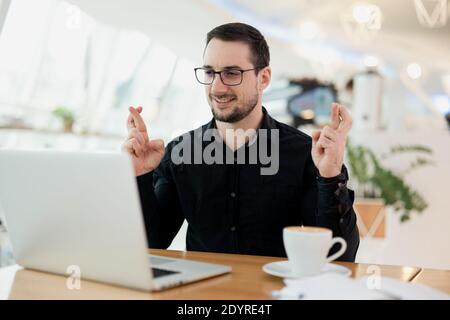 Mann in Brillen und schwarzem Hemd gestikulierenden Finger gekreuzt und lächelnd. Glück und abergläubisches Konzept. Junge attraktive Mann mit Bart sitzen in Stockfoto