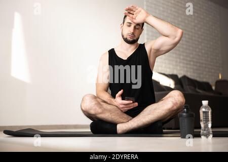 Muskulöser Mann wischt Schweiß von seiner Stirn nach hartem Hause Training. Gesundes Lifestyle-Konzept. Mann müde nach intensivem Training. Wasserflasche aus Kunststoff Stockfoto