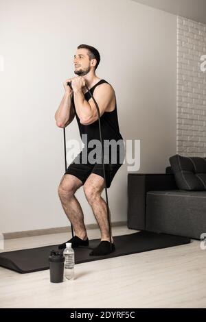 Fit Mann führt Übungen mit einem Widerstand Band. Hartes Training zu Hause. Minimalistisches Interieur im Hintergrund. Wasserflasche in der Nähe der Yogamatte. Ein Mann bekommt Stockfoto