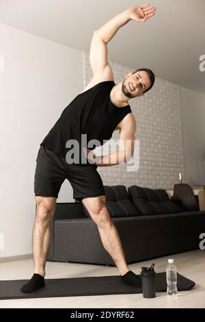 Fröhlicher junger Mann, der sich aufwärmt. Er streckt seine Arme, lächelt und schaut in die Kamera. Morgendliches Workout auf Yogamatte. Schwarze Sportswear. Starker Mann Stockfoto