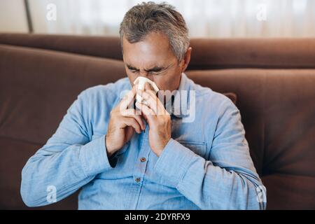 Kranker Mann mit laufender Nase isoliert sich zu Hause. Bleib zu Hause Konzept. Erwachsener Mann bläst sich die Nase, sitzt auf dem Sofa im Wohnzimmer. Isolieren Sie sich selbst, wenn Sie Stockfoto