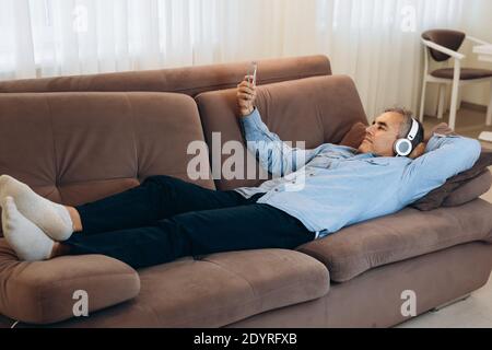 Ruhe zu Hause nach der Arbeit während der Quarantäne. Reifer Mann mit grauem Haar liegt auf dem Sofa und hört Musik in seinem Smartphone. Freizeit. Modern Stockfoto