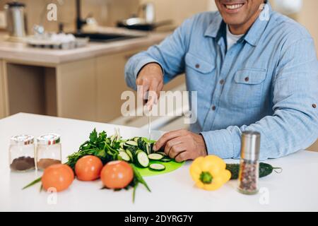 Erwachsene Mann zerkleinert Gemüse und Gemüse auf einem Küchentisch. Schnittansicht. Gurken, Tomaten, Paprika, Zucchinis und Gewürzgläser auf sauberem weißen Tisch Stockfoto