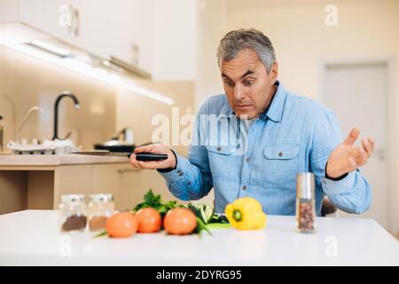 Amateur Koch Konzept. Überrascht reifer Mann weiß nicht, wie man ein Gericht kochen. Älterer Mann in blauem Hemd wirft seine Hände mit staunenden Blick auf seine Stockfoto